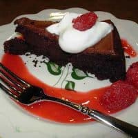 A slice of chocolate cake topped with whipped cream and a raspberry, served on a plate drizzled with rich raspberry sauce and garnished with two raspberries, next to a fork.
