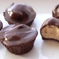 Four chocolate cups are shown on a white surface. One, resembling Chocolate Dipped Peanut Butter Balls, is bitten in half, revealing a creamy, light brown filling inside. The glossy chocolate coating is smooth and thick.