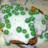 Two breaded cutlets topped with rich white gravy creamed peas, served on a speckled plate.