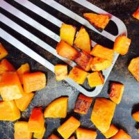 Diced roasted butternut squash pieces are being lifted from a dark baking tray with a metal spatula, showcasing the caramelized edges of the perfectly roasted butternut squash.