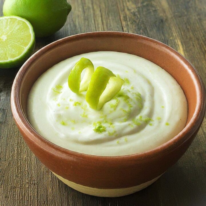 A brown bowl filled with creamy South Beach lime ricotta creme, garnished with lime zest and twisted lime peel. Halved and whole limes are in the background on a wooden surface.