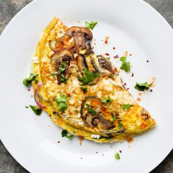 An asparagus mushroom omelet garnished with chopped herbs and red pepper flakes is served on a white plate. The omelet is folded in half, revealing slices of mushrooms, tender asparagus, and green herbs inside and on top.