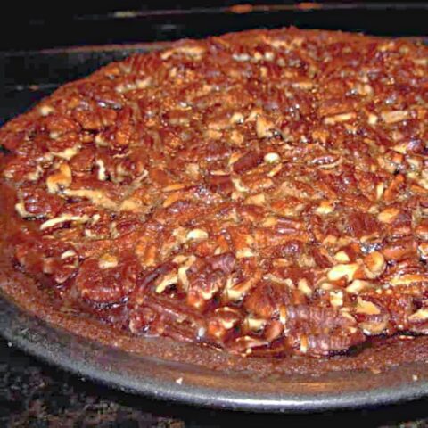 Close-up of a freshly baked Molasses Pumpkin Pecan Pie with a glossy, caramelized nut topping and a golden-brown crust, displayed on a dark baking pan.