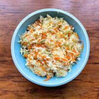 A blue bowl filled with shredded cabbage pineapple coleslaw, made of cabbage, carrots, and juicy pineapple, sits on a wooden surface.