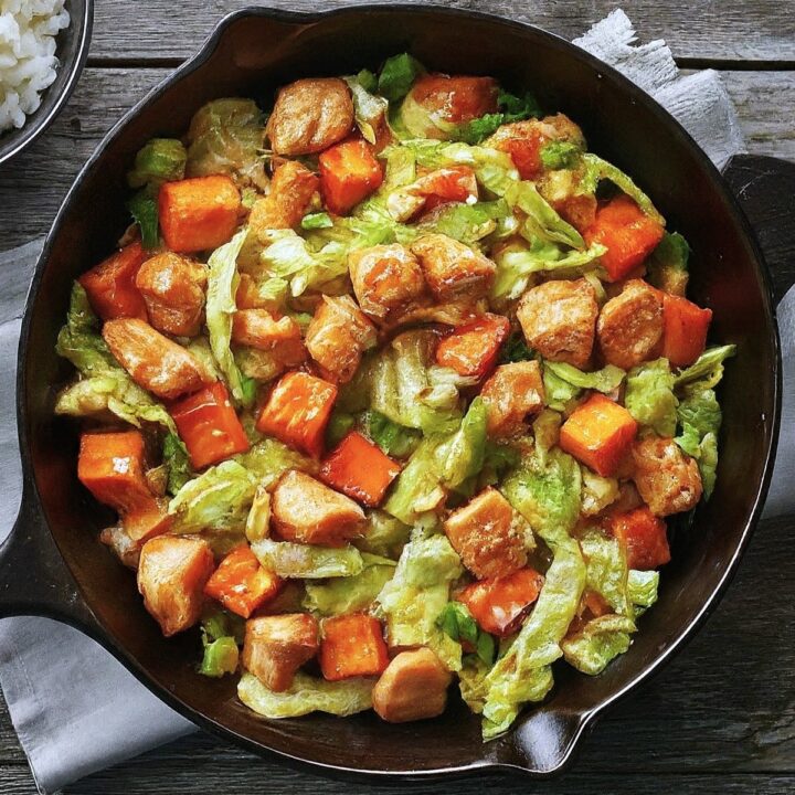 A skillet filled with Chicken Cabbage Sweet Potato Stir Fry, featuring tender chicken, chopped carrots, sweet potatoes, and cabbage in a savory sauce. The dish rests on a rustic wooden table with a bowl of white rice nearby.