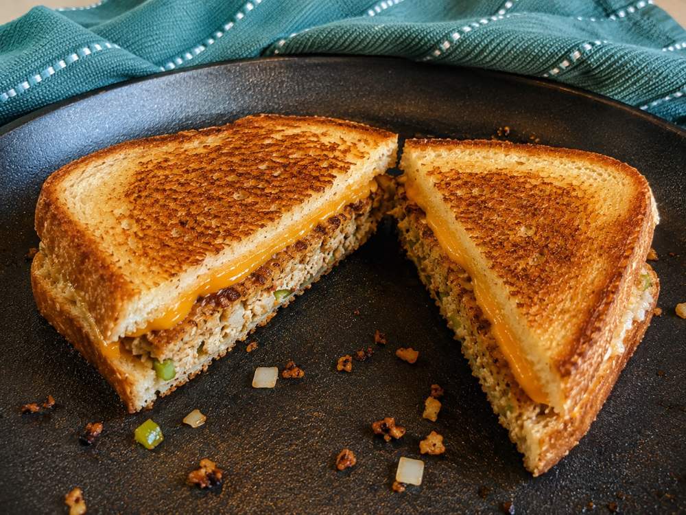 A grilled cheese sandwich with golden brown bread and melted cheese, cut in half on a black plate with diced onions and green peppers, sits next to a teal cloth—perfectly paired with homemade salmon patties for a comforting meal.