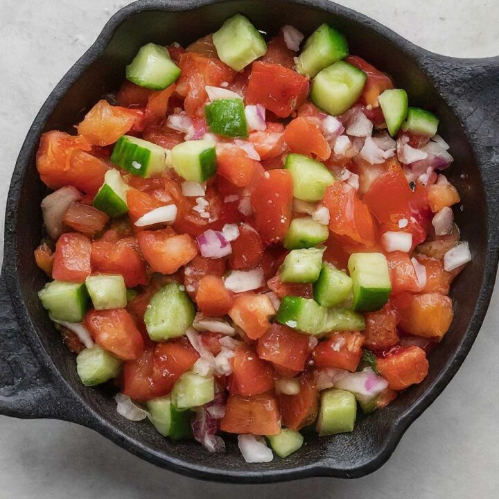 A black bowl filled with a fresh cucumber salsa featuring diced tomatoes, cucumbers, and red onions on a light-colored surface.