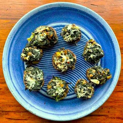 A blue plate with nine Spinach Stuffed Mushrooms topped with herbs and melted cheese, arranged in a loose circle on a wooden table.