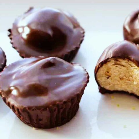 Chocolate peanut butter cups on a white surface, with one cup cut in half to reveal a creamy peanut butter filling. The glossy tops resemble Chocolate Dipped Peanut Butter Balls, making them even more tempting.