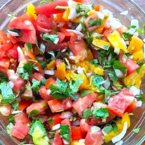 A glass bowl filled with Fresh Pico de Gallo—colorful salsa made of chopped tomatoes, yellow peppers, onions, and fresh cilantro—sits on a wooden surface.