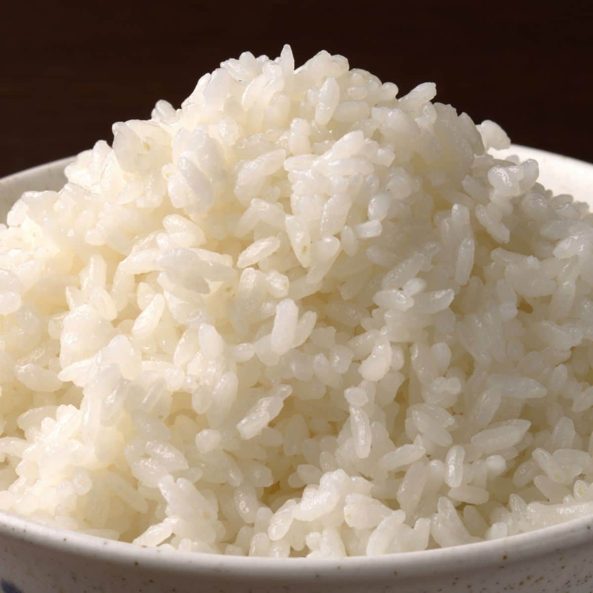 A close-up of a bowl filled with steaming, fluffy white rice, with individual grains visible and a soft, glossy texture.