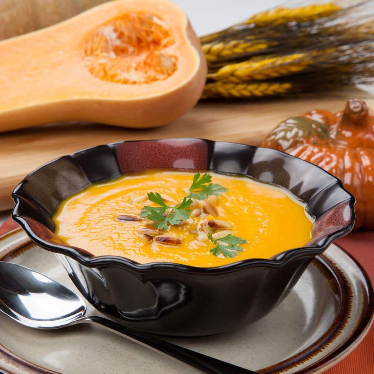 a gorgeous burgundy bowl of butternut squash soup on a plate, with a spoon and a pumpkin decoration next to it.