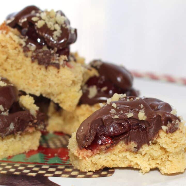 A close-up of crumbly Christmas cookie bars topped with chocolate frosting, cherry filling, and sprinkled nuts, stacked on a festive plate.