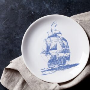 A white plate with a blue ship design, and sitting on a tan colored cloth napkin.