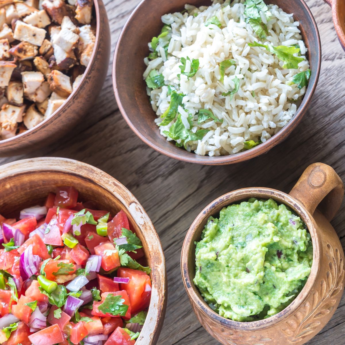 a few of the necessary burrito bowl ingredients: chicken, chopped vegetables, rice, guacamole, and more.