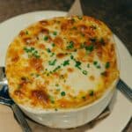 A bowl of French onion soup topped with melted, golden-brown cheese and sprinkled with chopped chives, served on a white plate with a spoon beside it—perfect for those curious about how to eat French onion soup properly.