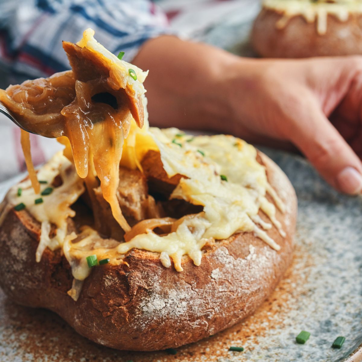 a man's hand taking a spoonful of French onion soup from a bread bowl.