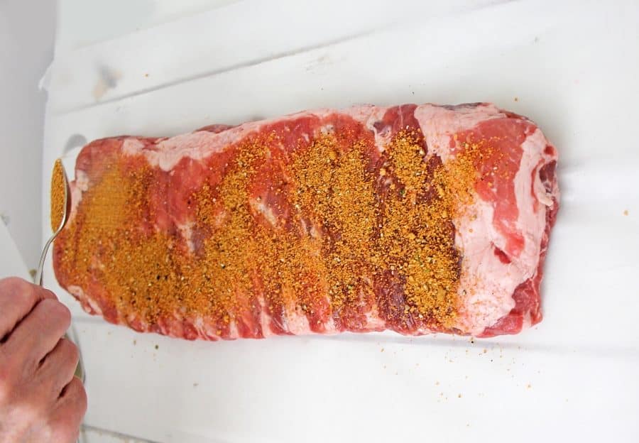 A hand sprinkles a generous amount of dry seasoning rub onto a rack of raw pork ribs placed on a white surface, prepping them for easy Instant Pot Dump and go Country Style Ribs.