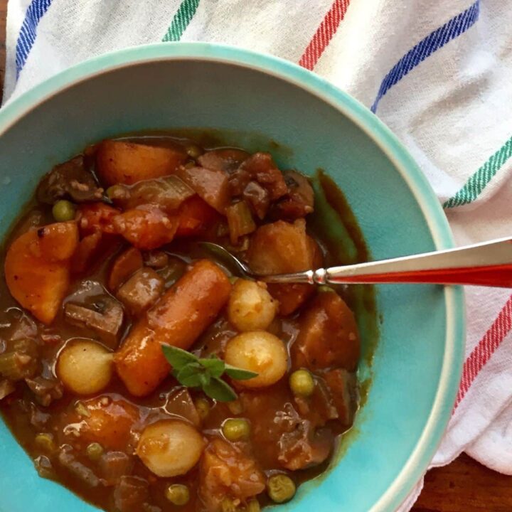 A bowl of hearty Instant Pot Vegetarian Bourguignon stew with carrots, potatoes, peas, and pearl onions, garnished with a small sprig of herbs. A spoon rests in the bowl, and a striped cloth is in the background.