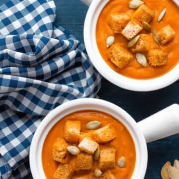 Two bowls of creamy pumpkin soup topped with crunchy croutons and pumpkin seeds.