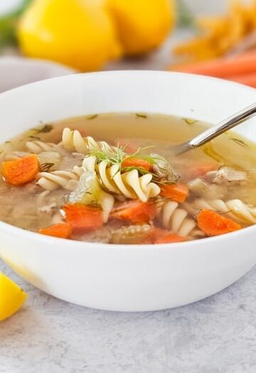 A white bowl of chicken noodle soup with carrots, rotini pasta, and herbs sits on a light surface. A spoon rests in the bowl, and a lemon wedge and dill garnish are beside it. Blurred lemons and carrots are in the background.