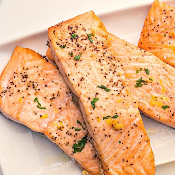 Three South Beach Diet Grilled Salmon fillets, seasoned with black pepper, herbs, and lemon zest, are arranged on a white rectangular plate. The salmon has a light golden color and is garnished with chopped parsley.