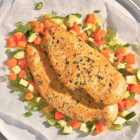 Two pieces of seasoned, cooked South Beach Chicken en Papillote rest on parchment paper atop a bed of chopped vegetables, including carrots, zucchini, and green onions, served on a white plate.