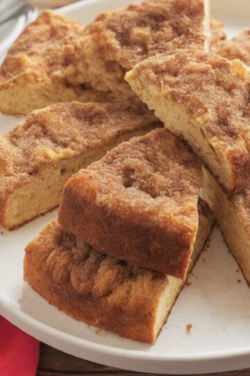 Slices of Snickerdoodle Coffee Cake arranged in a circle on a white plate, showing a golden-brown crumb topping. A red napkin and stacked plates with forks are visible in the background.