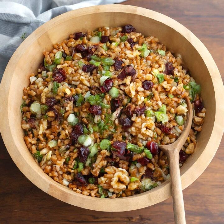A wooden bowl filled with a mixed winter wheat berry salad of cooked brown rice, green onions, dried cranberries, and chopped nuts, with a wooden serving spoon resting on the side.
