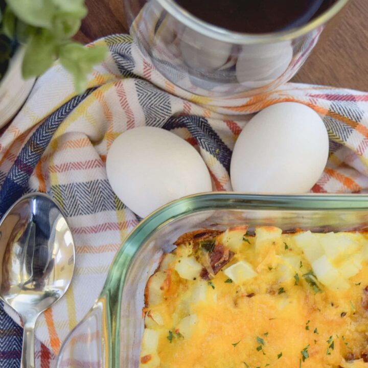 A glass baking dish filled with cheesy hash brown breakfast casserole sits next to two whole eggs, a spoon, a glass of dark coffee, and a plaid napkin on a wooden table.