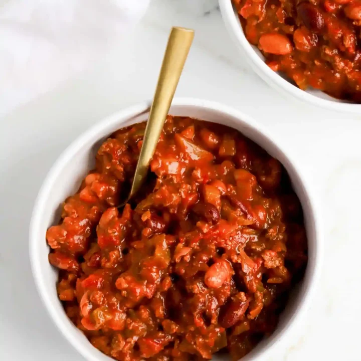Two white bowls filled with thick, chunky chili made with beans and tomatoes—a Childhood Comfort Foods classic. A gold spoon rests in one bowl, all set on a white marble surface with a white napkin nearby.