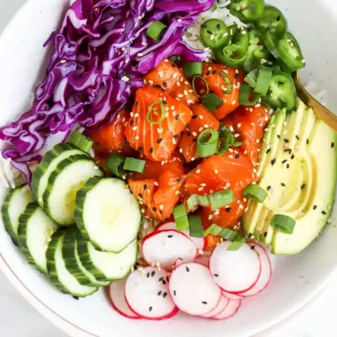 A colorful poke bowl topped with cubed salmon, sliced cucumber, radish, avocado, jalapeño, shredded purple cabbage, green onions, and sprinkled with sesame seeds—perfect inspiration for your next Salmon Recipes creation.