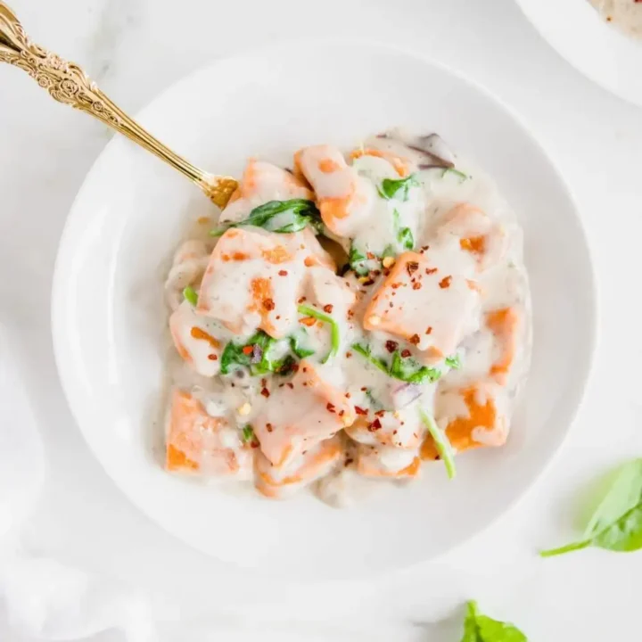 A white bowl filled with creamy gnocchi, spinach, and a white sauce, garnished with red pepper flakes. A gold spoon rests in the bowl. Fresh spinach leaves are scattered on the white surface—a cozy pick for Sweet Potato Recipes lovers.