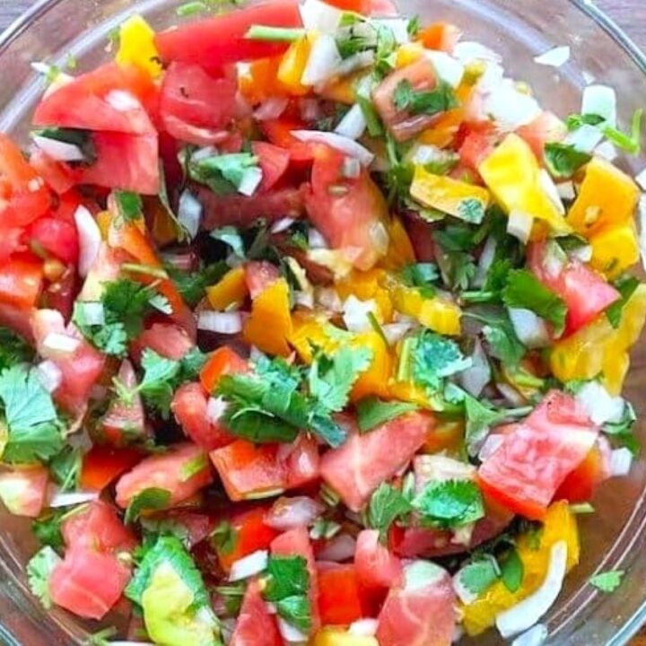 A glass bowl filled with Fresh Pico de Gallo made of chopped tomatoes, yellow peppers, onions, cilantro, and herbs sits on a wooden surface.