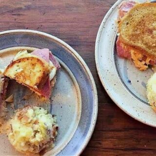 Two ceramic plates each hold a grilled sandwich, inspired by a Corned Beef Reuben Sandwich Recipe, filled with ham and cheese, plus potato salad, all atop a wooden surface.