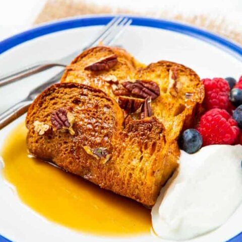 Two slices of French toast, inspired by classic breakfast casseroles, topped with pecans and served with maple syrup, whipped cream, fresh raspberries, and blueberries on a white plate with a blue rim.