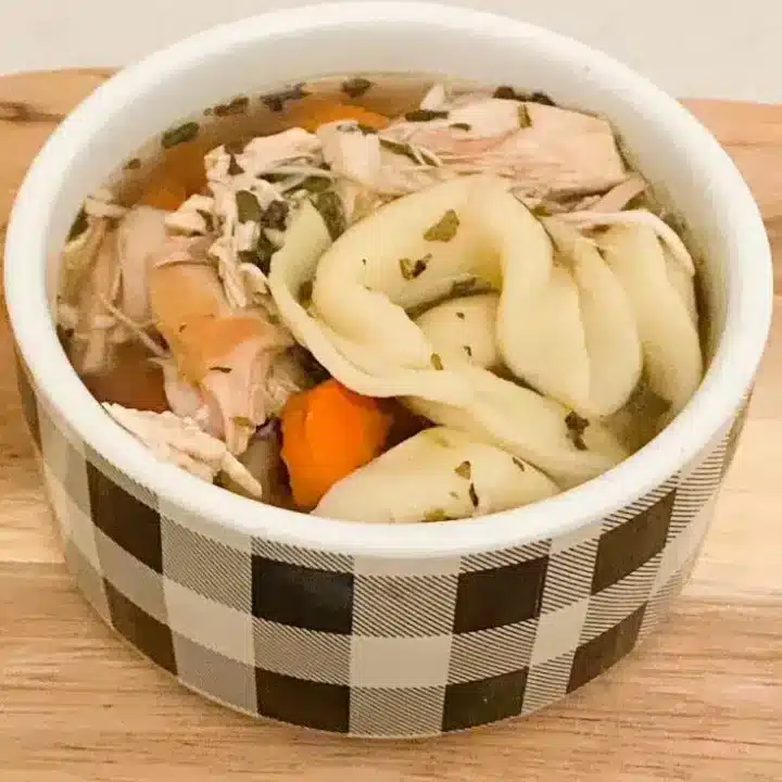 A black and white checkered bowl filled with soup featuring tortellini pasta, shredded chicken, carrots, and herbs sits on a wooden surface—a cozy twist on classic chicken soup recipes.
