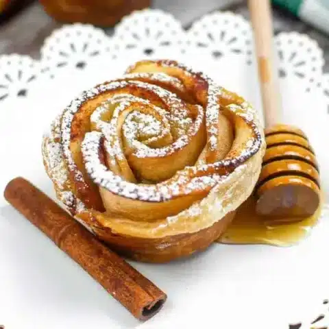 A rose-shaped pastry, perfect for fall desserts, is dusted with powdered sugar and sits on a white doily plate beside a cinnamon stick and a honey dipper drizzled with honey.