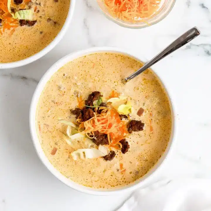 A bowl of creamy Fall Soup with ground meat, shredded cheese, and cabbage on top sits invitingly with a spoon inside. Another bowl and a small dish of shredded cheese rest nearby on the white marble surface.