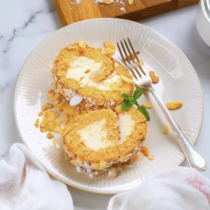 Two slices of a cream-filled Swiss roll, perfect for Fall Desserts, are garnished with sliced almonds on a white plate with a fork. A sprig of mint and a white cloth sit nearby, with a cutting board and more almonds in the background.