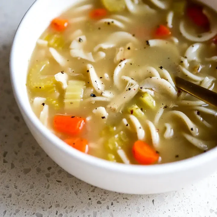 A white bowl filled with noodle soup, inspired by classic Chicken Soup Recipes, features sliced carrots, celery, noodles, and a light broth, placed on a speckled countertop.