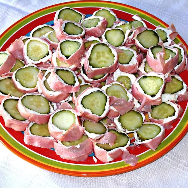 A colorful plate filled with pickle roll-ups—sliced cucumber pieces wrapped in ham and a layer of soft white cheese—arranged neatly on a white tablecloth.