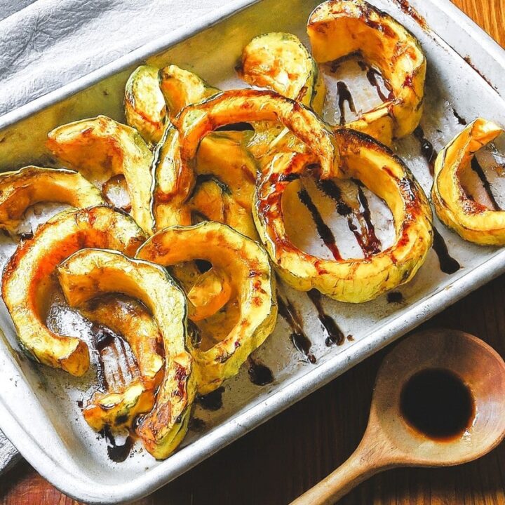 Slices of Two Ingredient Roasted Sweet Dumpling Squash arranged on a baking sheet, drizzled with balsamic glaze. A wooden spoon with extra glaze rests nearby on a wooden table.