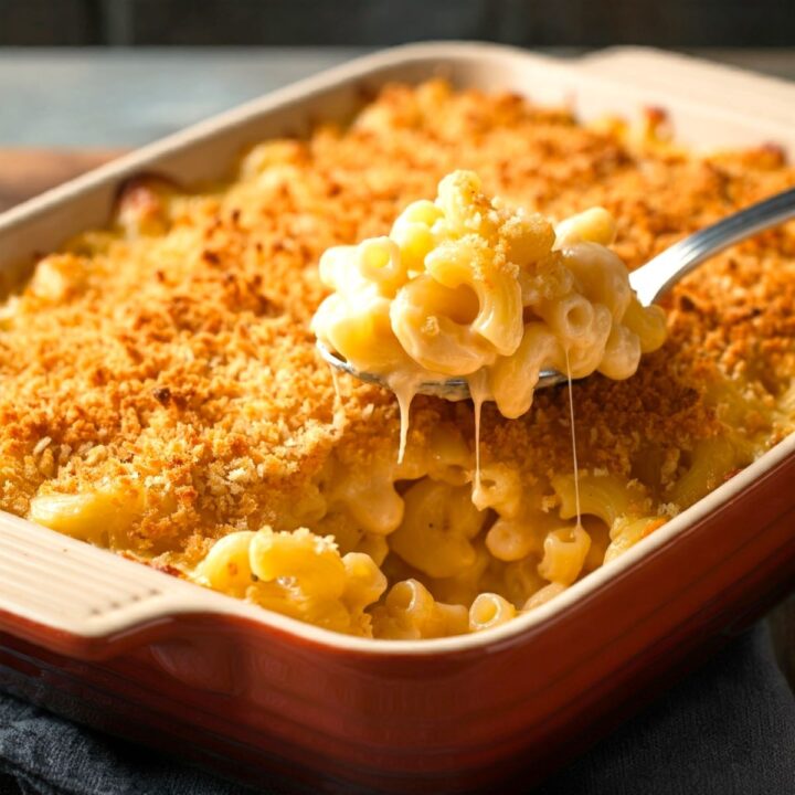 A close-up of baked macaroni and cheese in a rectangular dish, with a spoon lifting a serving showing creamy, melted cheese and a golden breadcrumb topping.