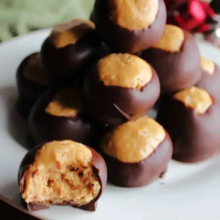 A plate stacked with chocolate-covered peanut butter balls—candy made with cereal—one in front with a bite taken out, revealing creamy peanut butter filling inside. Some red and silver ornaments are blurred in the background.