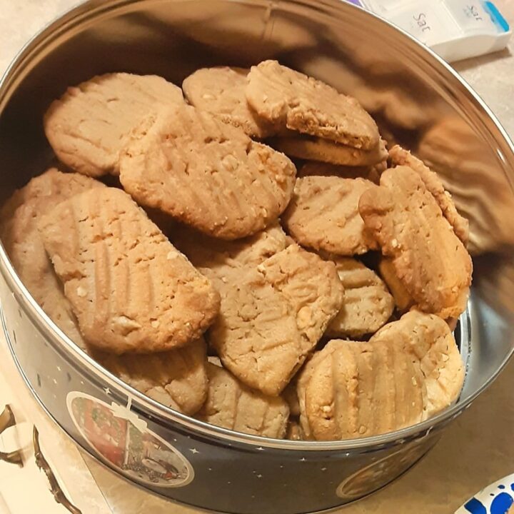 A round metal tin filled with the best crunchy peanut butter cookies, each marked with a classic crisscross fork pattern, sits open on a countertop.