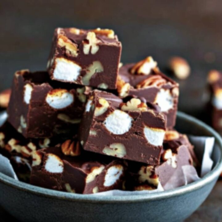 A bowl filled with squares of Rocky Road Fudge, each piece containing visible marshmallows and pecan nuts, stacked on top of each other against a dark background.