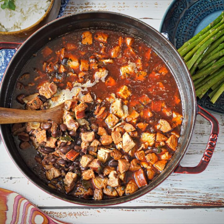 A pot of stew with cubed meat, beans, and vegetables in a rich sauce is stirred with a wooden spoon—one of the hearty Recipes Made with Dried Beef. Sides of white rice and roasted asparagus are served on plates nearby.