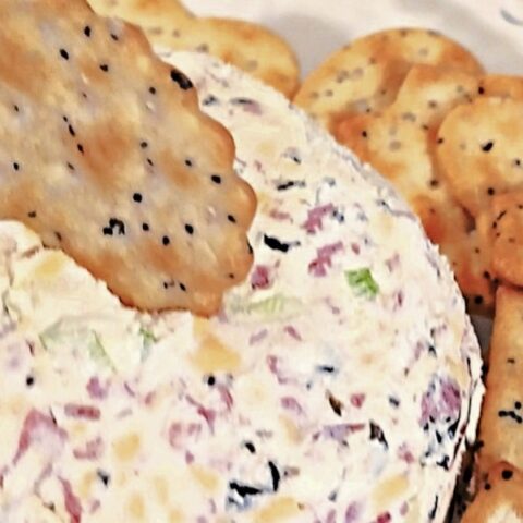 A close-up of a dried beef cream cheese ball mixed with herbs and bits of meat, surrounded by seeded crackers, with one cracker partially stuck into the cheese ball.