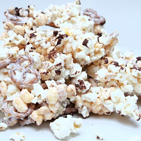A pile of almond bark popcorn clusters drizzled with chocolate and coated with white icing sits on a white surface. Some pieces appear to have pretzels mixed in, adding extra crunch to the sweet treat.